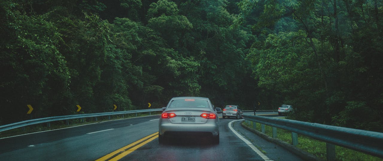 Cars driving on a winding forest road surrounded by lush greenery and overcast sky.