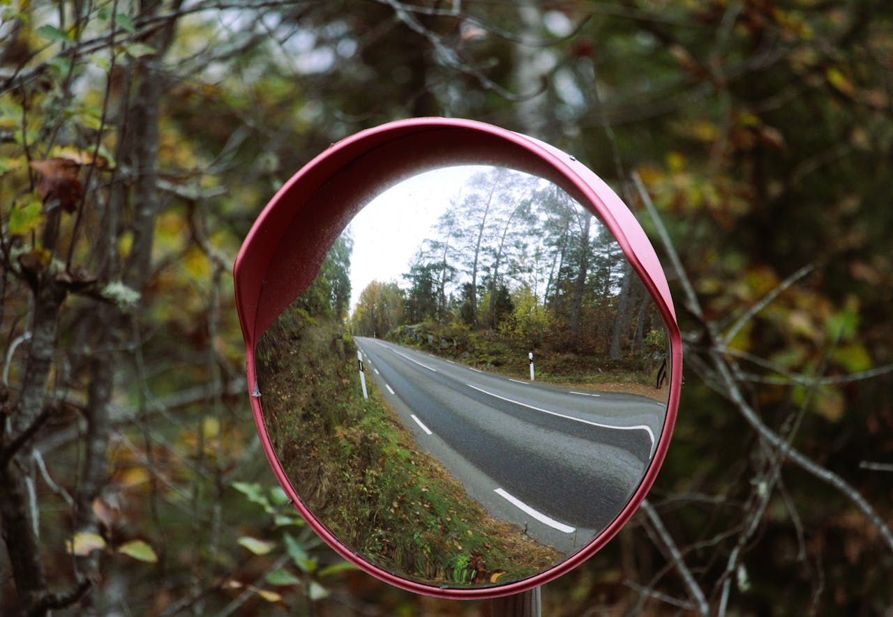 A scenic autumn road captured through a red safety mirror with vibrant fall foliage.